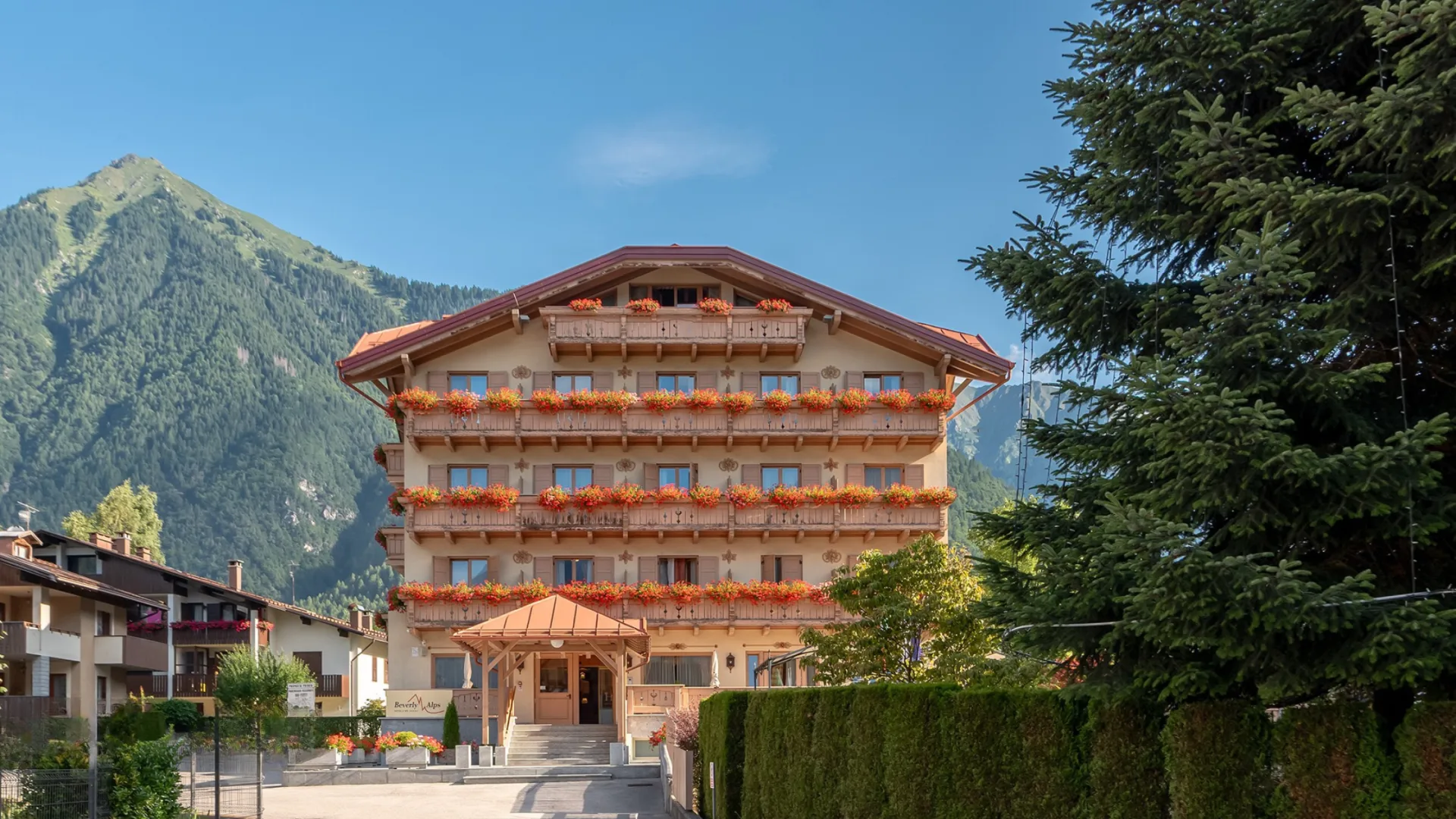 Facciata estiva dell'hotel con balconi fioriti di rosso e montagne verdi